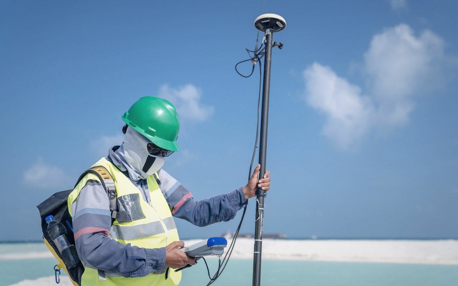 Surveyor in a hard hat holds a GPS pole while checking a handheld receiver near the shore